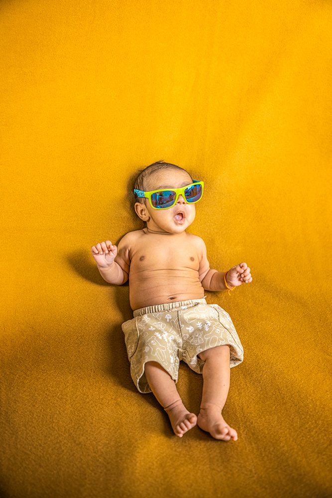 Baby wearing colorful sunglasses and beige tropical print swim shorts lies on bright orange background with arms outstretched