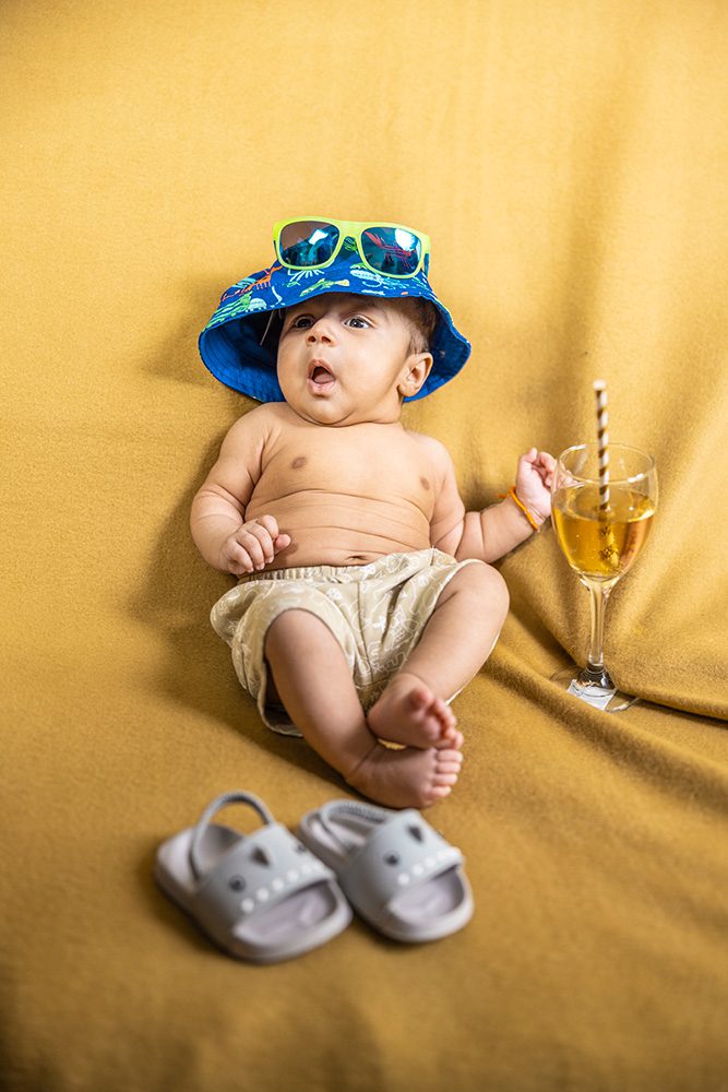 Baby wearing blue tropical bucket hat with sunglasses and light-colored shorts lies on yellow background next to champagne glass and small sandals