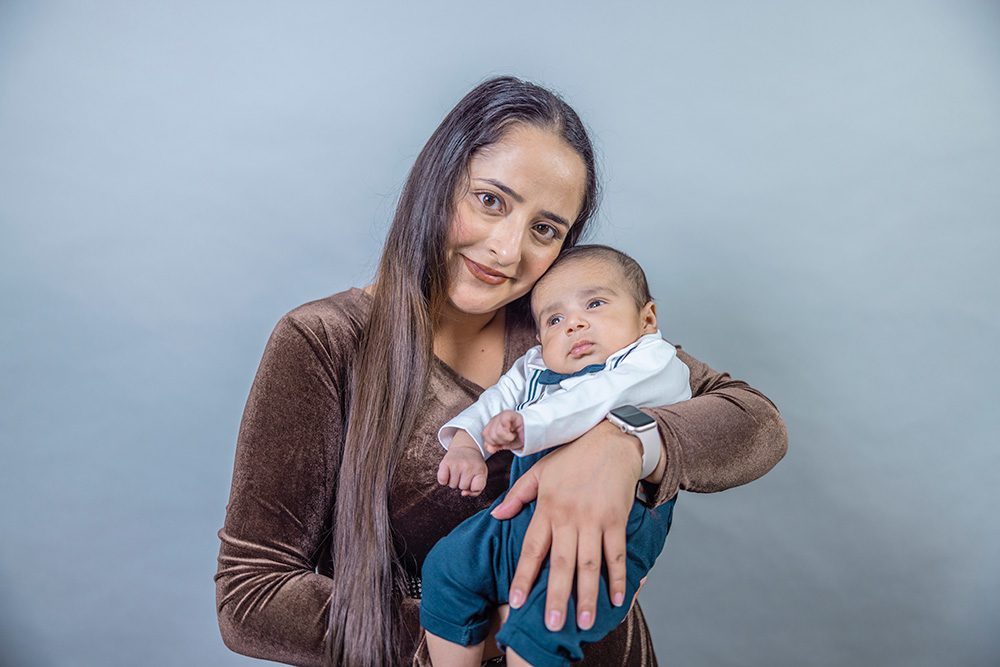 Young woman with long brown hair wearing a brown sweater holds a baby in white and blue clothing against a light blue background