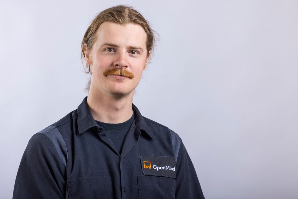 Professional headshot of a young man with a mustache and shoulder-length hair wearing a dark OpenMind work shirt against a light gray background