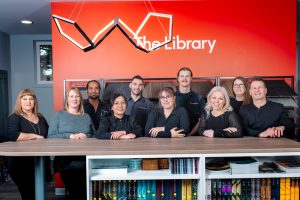 Professional team of ten people in business attire posing behind wooden counter at The Library with red accent wall and bookshelves below