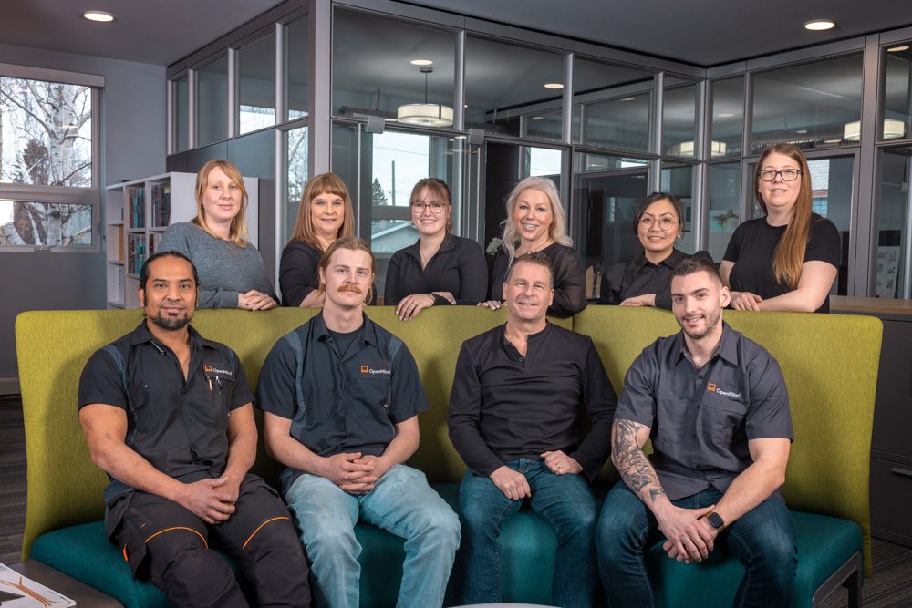 Ten-person team photo with four men seated on a green and yellow modular sofa and six women standing behind them in a modern office with glass walls