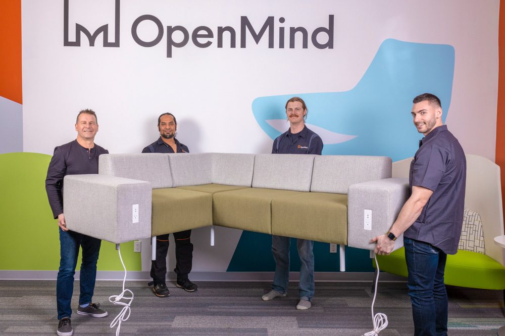 Alt Text: Four men in dark shirts holding a large modular office sofa with green and gray cushions in front of a wall displaying the OpenMind logo