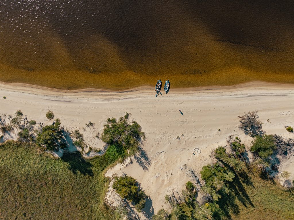 Aerial view of a pristine beach with two blue boats on sandy shore, surrounded by lush green vegetation and calm brown-tinted water