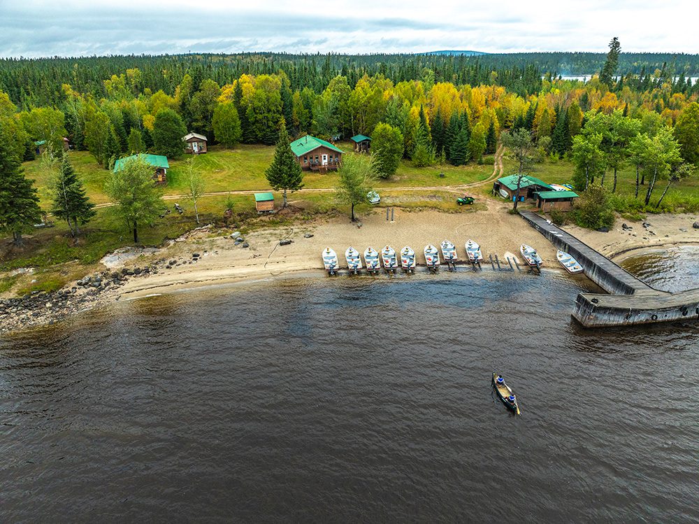 Aerial view of wilderness fishing lodge with green-roofed cabins, wooden dock, fleet of white boats, and autumn forest surrounding a lake