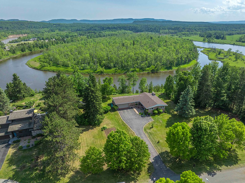 Aerial view of waterfront property with ranch-style home, curved driveway, and expansive lawn overlooking a winding river surrounded by dense forest and rolling hills