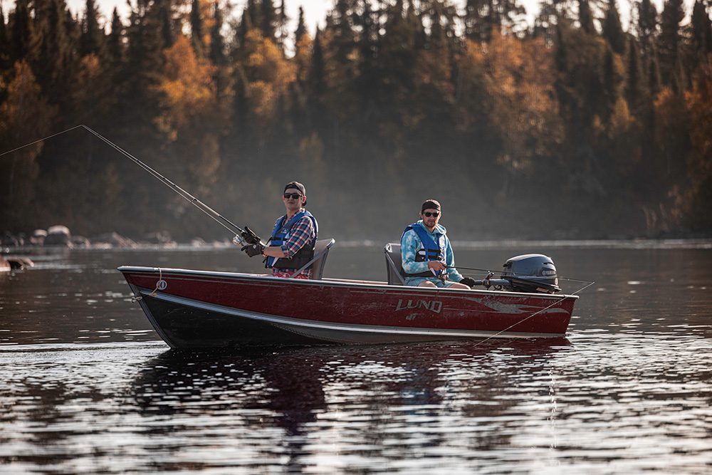 Two men fishing from red Lund boat on calm lake during golden hour with autumn forest and mist in background