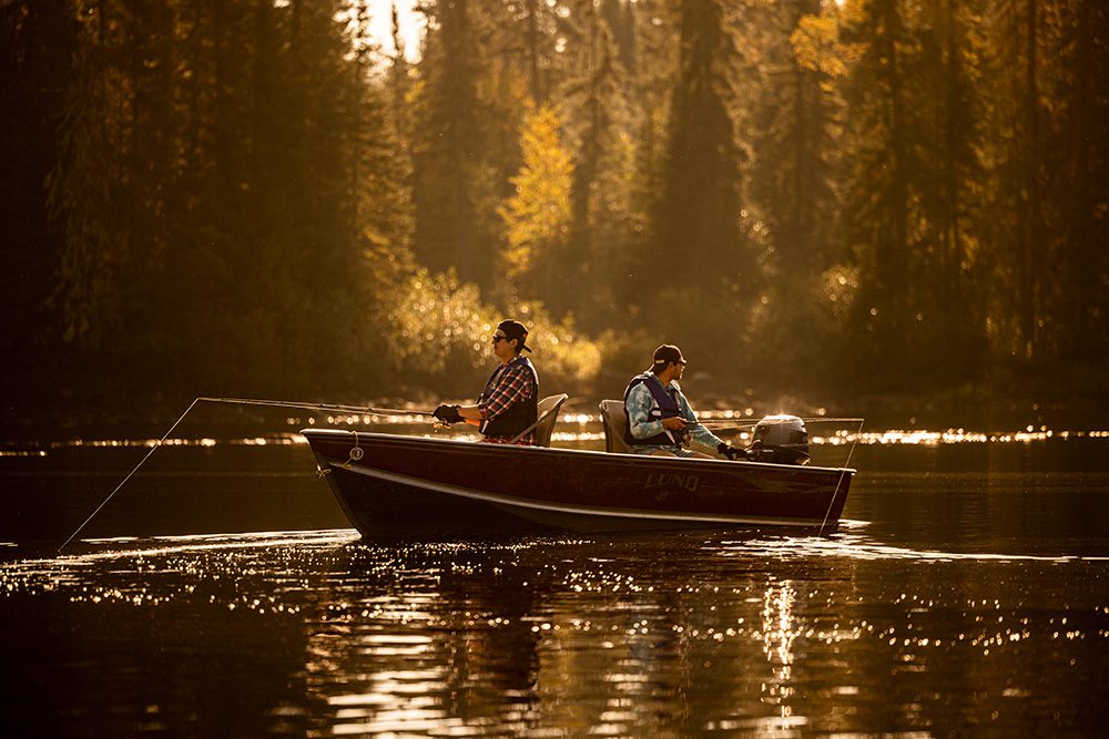Two men fishing from red Lund boat on calm lake during golden hour with autumn forest and mist in background