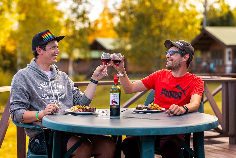 Two men toasting with wine glasses while dining at outdoor table with autumn foliage and cabin in background
