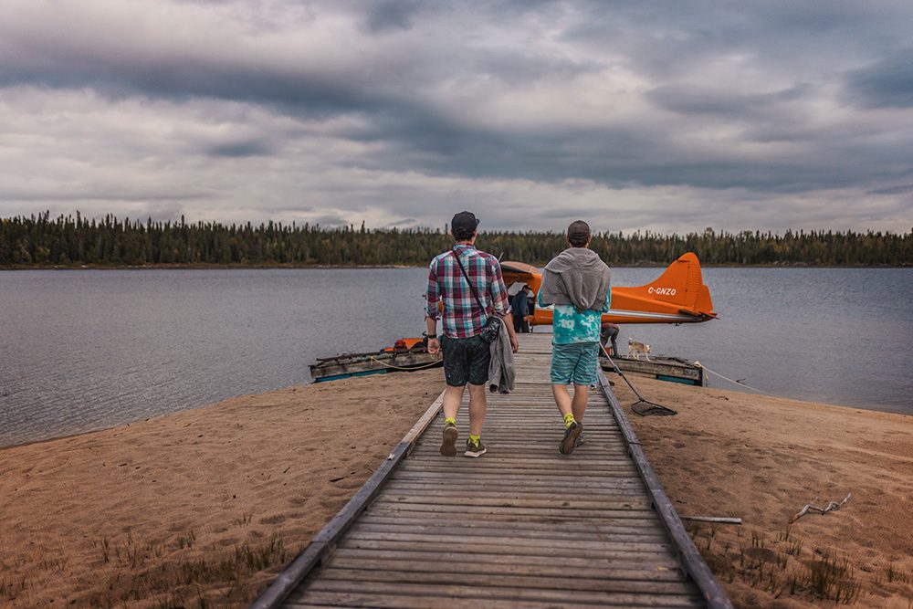 Two men walking on wooden dock toward orange seaplane with wilderness lake and forest under cloudy sky
