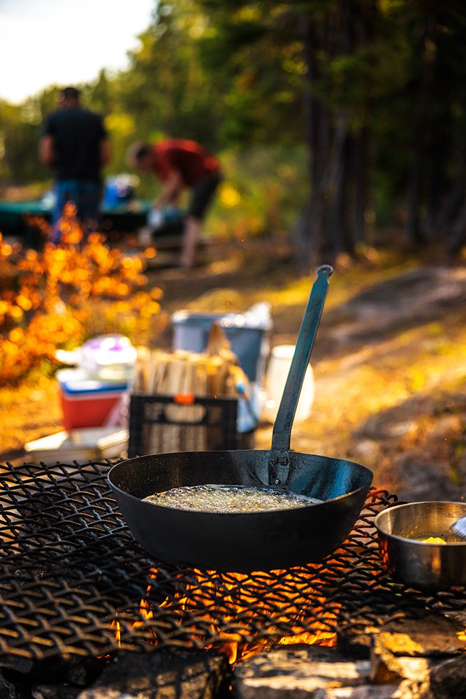Cast iron skillet cooking over campfire with two people preparing food in blurred background during golden hour