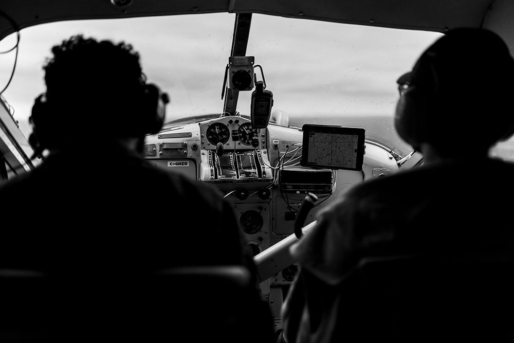 Black and white photo taken from inside small aircraft cockpit showing two passengers and pilot with instrument panel and GPS screen visible
