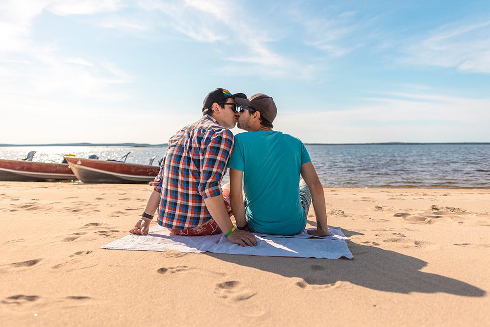 A couple sitting on a beach towel sharing a kiss while facing the water on a sunny day.