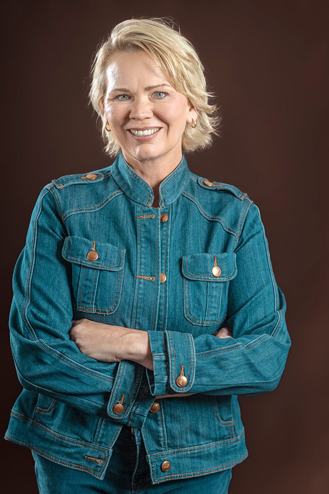 Professional headshot of a woman with short blonde hair wearing a denim jacket with arms crossed against a brown background