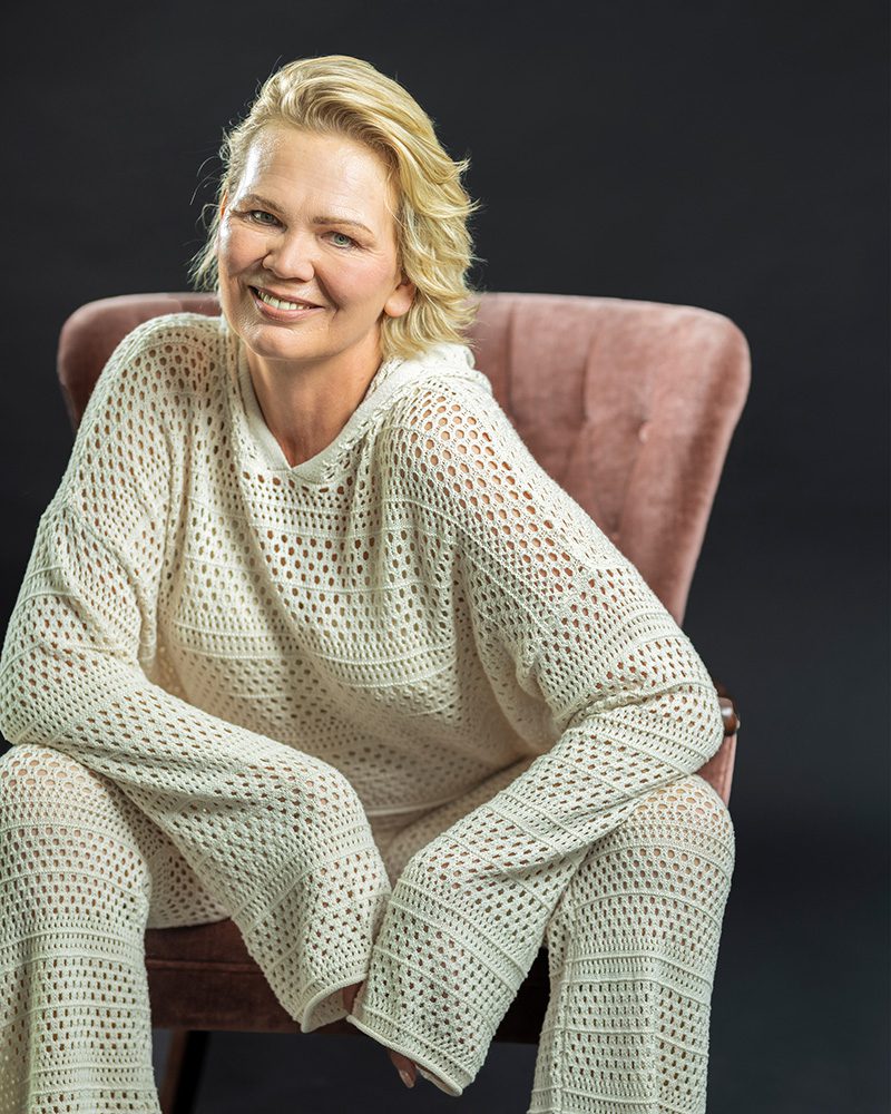 Professional portrait of a woman with short blonde hair wearing a cream-colored open-weave knit outfit while sitting in a pink velvet chair against a dark background