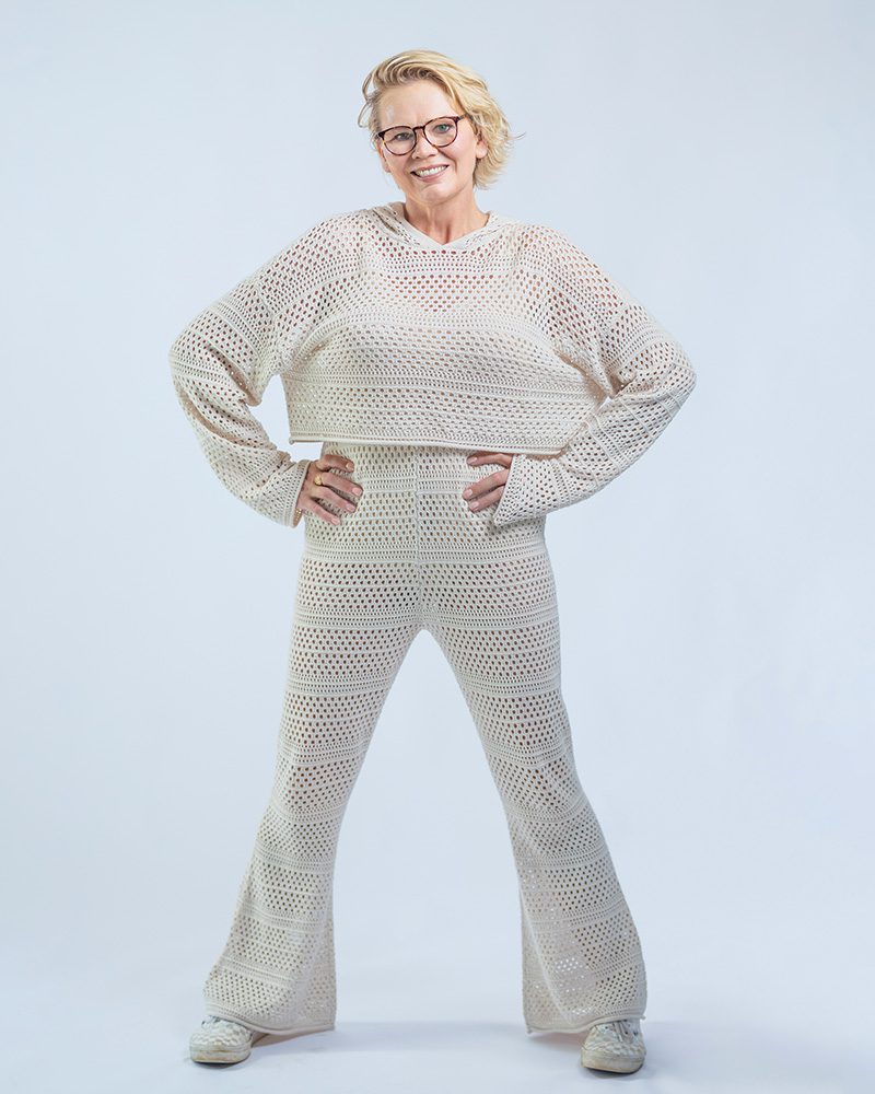 Professional portrait of a woman with short blonde hair and glasses wearing a beige knit outfit with hands on hips against a white background
