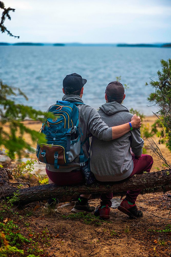 Two men sitting together on fallen log overlooking lake, one with arm around the other's shoulder, wearing hiking gear and backpacks