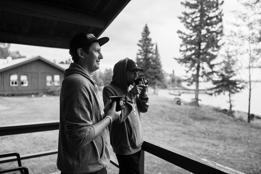 Black and white photo of two men holding coffee mugs on covered porch overlooking snowy landscape with cabin and evergreen trees