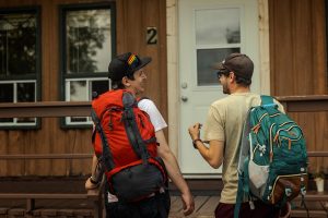 Two men with hiking backpacks standing on wooden cabin porch, one unlocking door marked with number 2