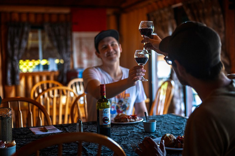 A couple clinking glasses of red wine across a dining table inside a cozy wooden cabin.