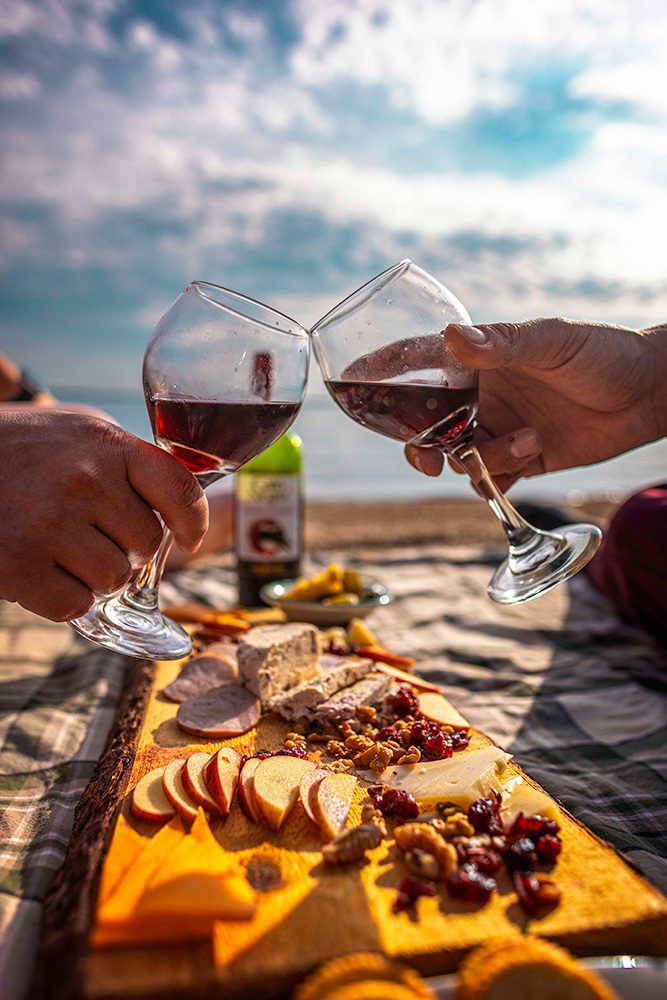 A couple clinks glasses of red wine over a picnic spread with cheese, fruits, and crackers on a beach under a partly cloudy sky.