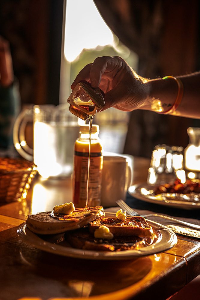 A hand pouring syrup onto a stack of pancakes topped with butter, with warm sunlight streaming through a window in the background.