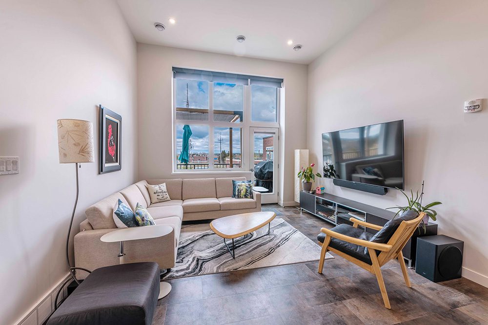 Modern living room with beige sectional sofa, mid-century wood chair, oval coffee table, large TV, and floor-to-ceiling windows overlooking urban views