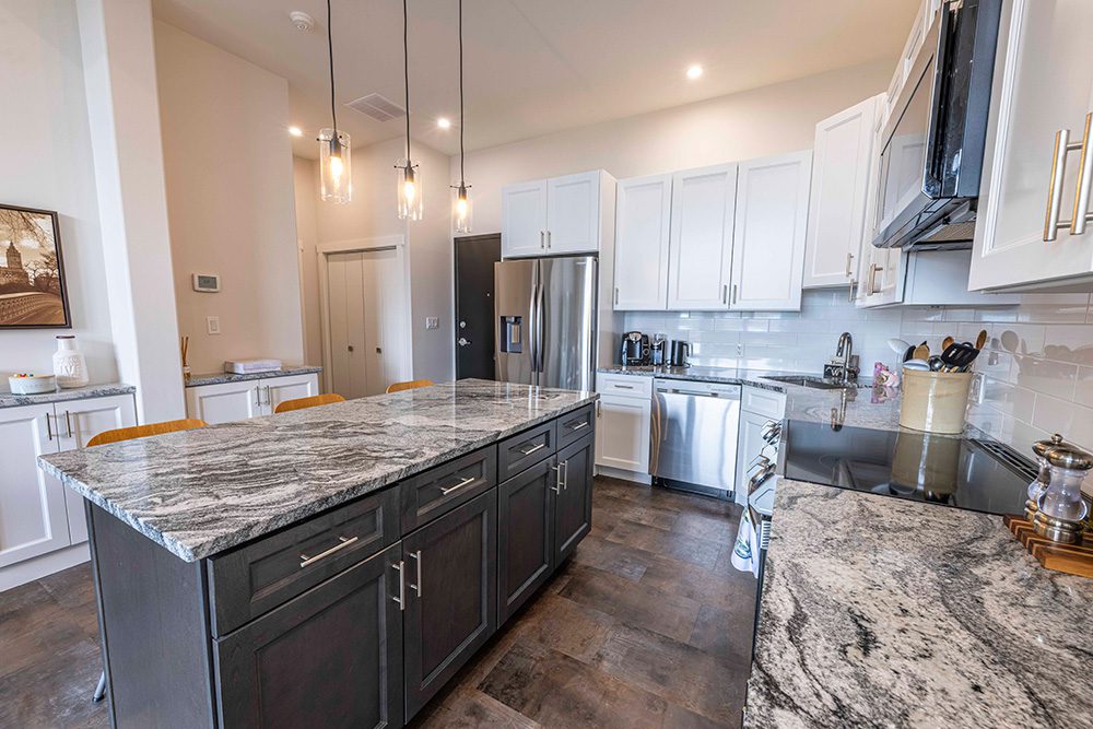 Luxurious kitchen with white upper cabinets, dark gray island, dramatic granite countertops, stainless steel appliances, and glass pendant lights