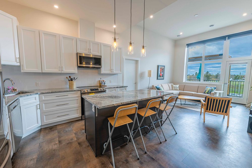 Modern open-concept kitchen with white cabinets, dark island, granite countertops, pendant lights, and connected living area with large windows