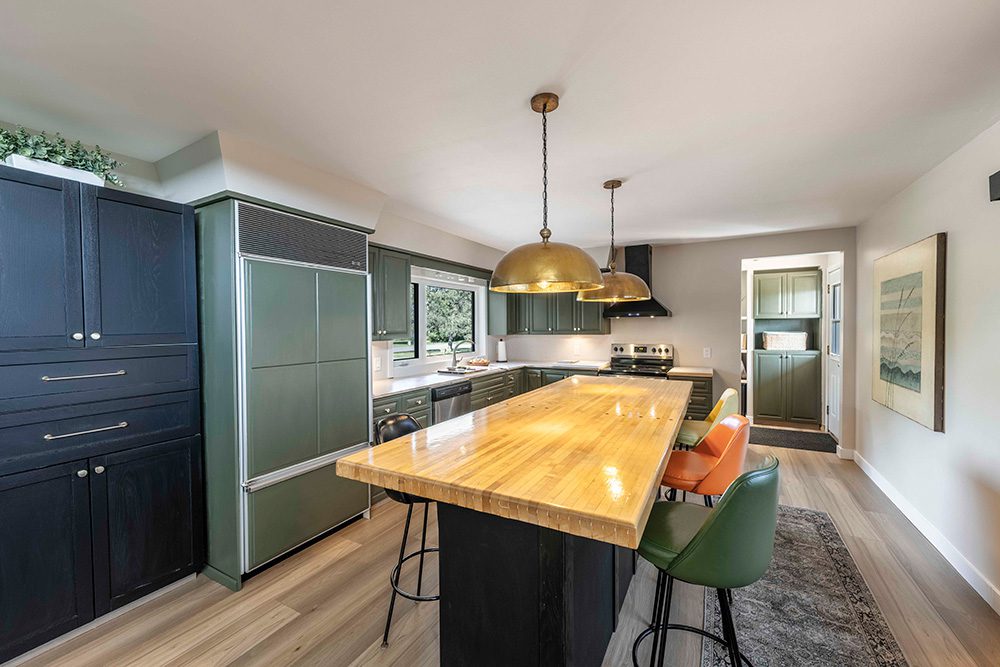 Modern kitchen with sage green cabinets, butcher block island, brass pendant lights, and colorful bar stools on hardwood floors