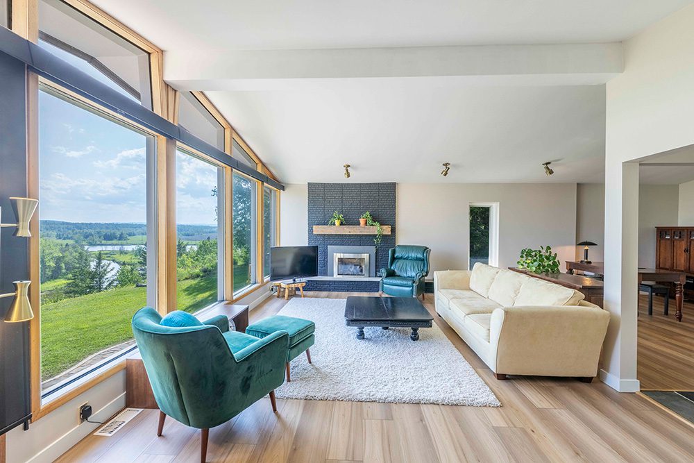 Modern living room with floor-to-ceiling windows, teal accent chairs, white sectional sofa, and brick fireplace overlooking scenic countryside views
