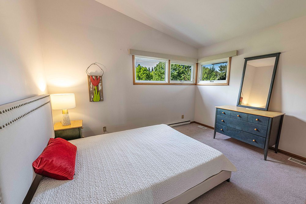 Clean modern bedroom with platform bed, white bedding, red accent pillow, blue dresser with mirror, and three windows showing green trees outside
