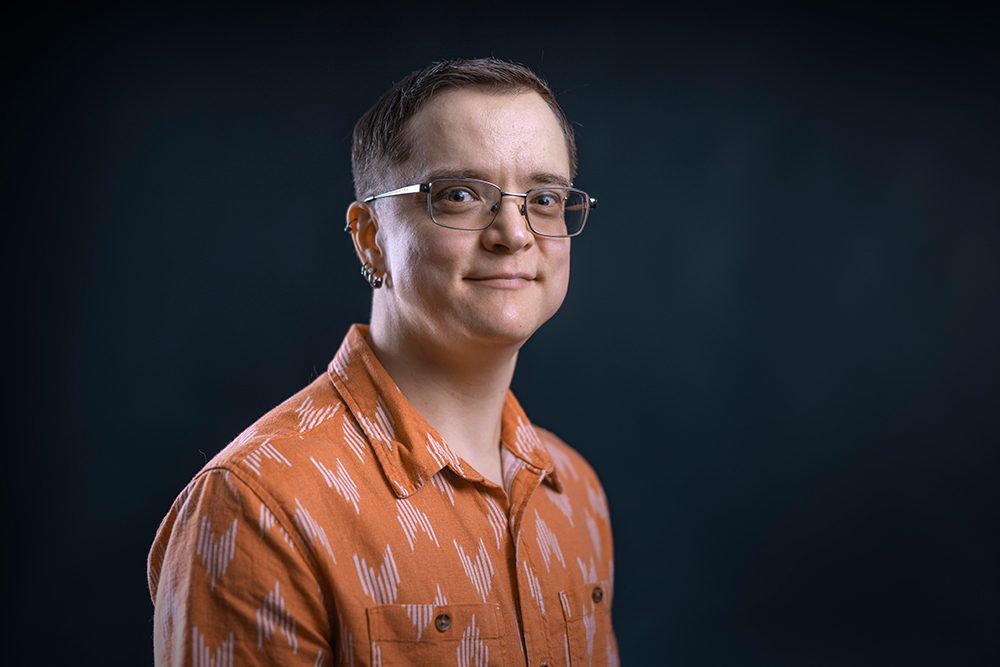 Professional headshot of a person with short brown hair and glasses wearing an orange patterned button-up shirt against a dark blue background