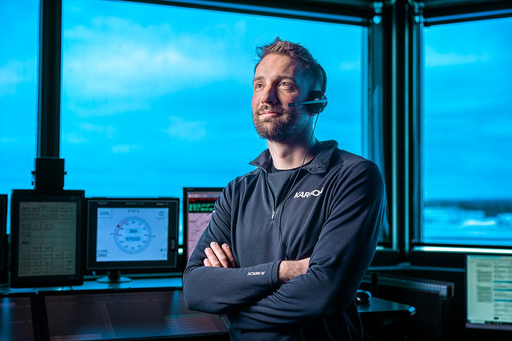Man wearing a headset in an air traffic control room with screens and large windows showing a blue sky.