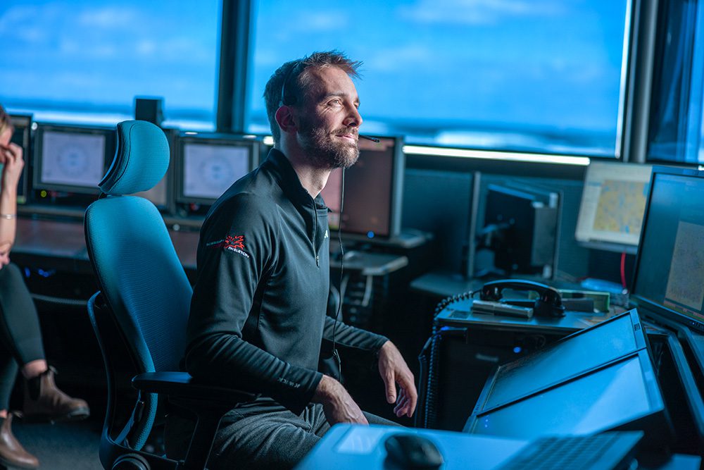 Man seated at a workstation in an air traffic control room, wearing a headset and looking up with a slight smile.