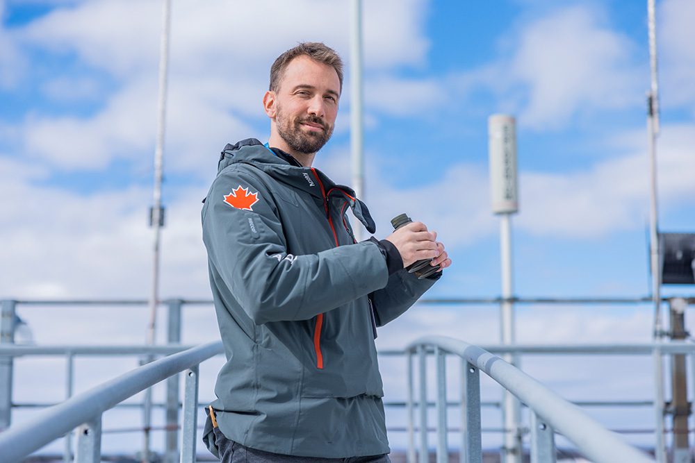 Man with beard wearing gray jacket with Canadian maple leaf patch holding water bottle on Thunder Bay Airport control tower observation deck with blue sky background