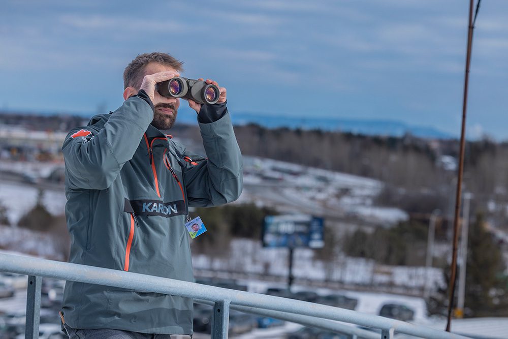 Man in gray flight suit with Canadian maple leaf patch looking through binoculars on observation deck with winter landscape and blue sky in background