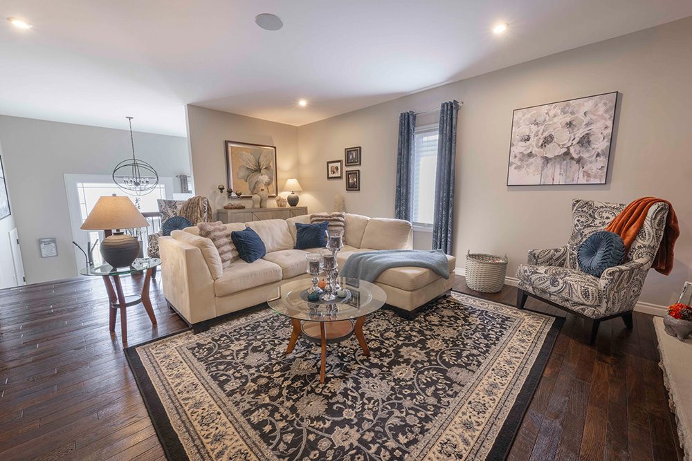 Open-concept living room with beige sectional sofa, patterned accent chair, glass coffee table, traditional area rug, and recessed lighting on dark hardwood floors