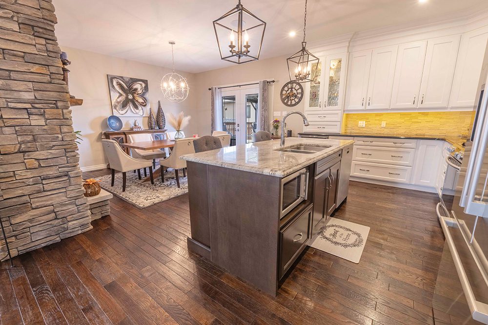 Open-concept kitchen and dining area with white cabinets, dark island, yellow subway tile backsplash, lantern pendant lights, and stone fireplace wall