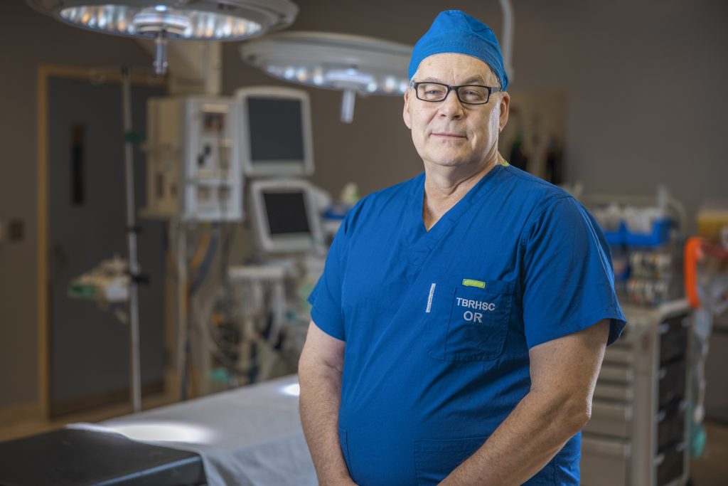 Surgeon in blue scrubs and cap standing confidently in a hospital operating room.