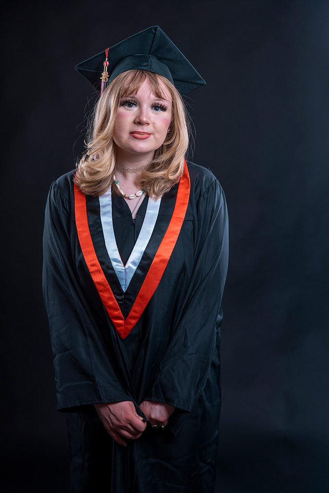 A young woman in a black graduation gown and cap, wearing a red, white, and black V-shaped stole, stands against a dark background.