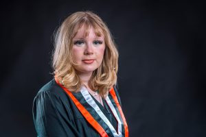 Young woman in black graduation cap and gown smiling at camera against dark gray background