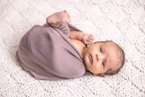Alert newborn baby wrapped in mauve blanket with one foot visible on white textured backdrop