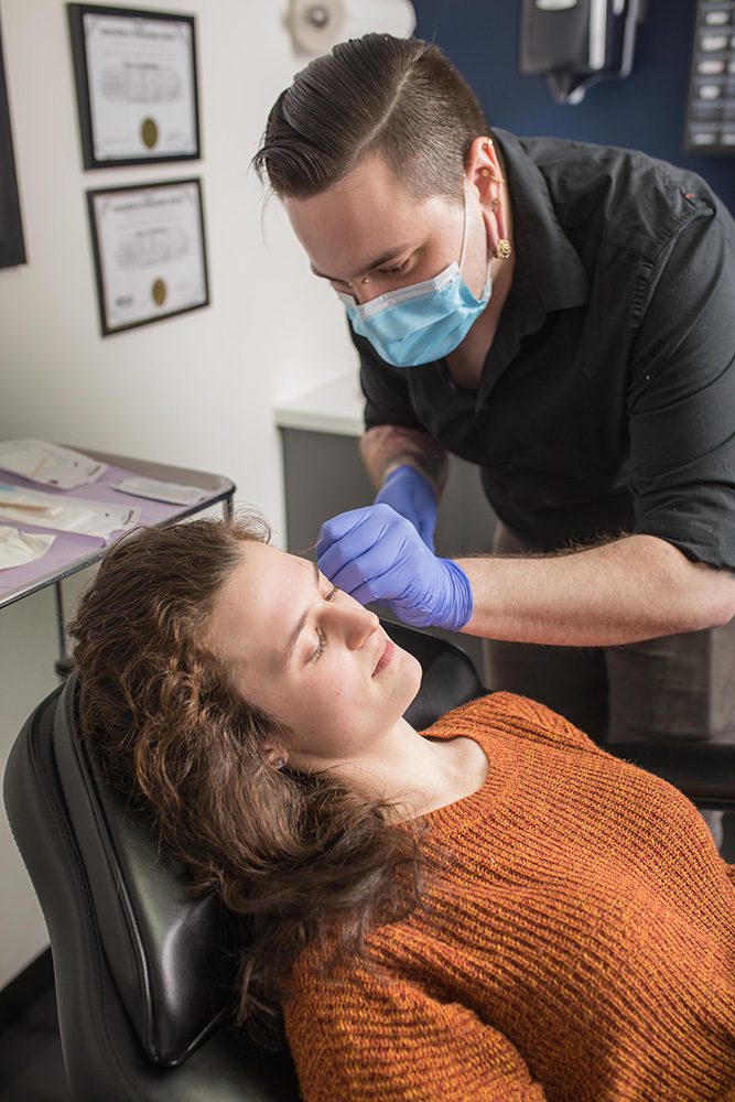 Professional performing facial piercing procedure on client in orange sweater at Identity Tattoo and Piercing studio