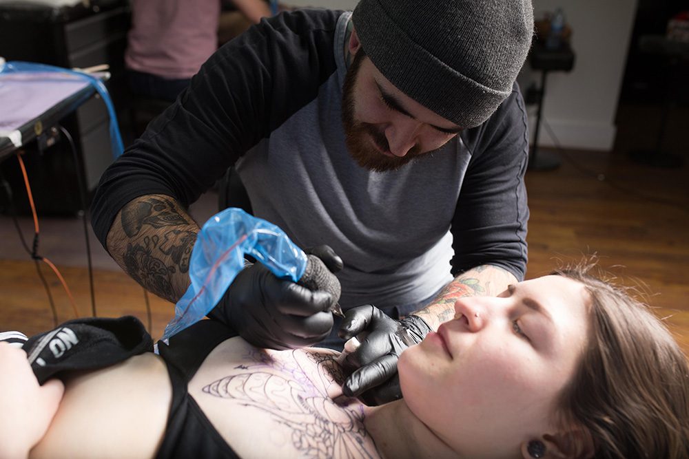 Tattoo artist creating detailed chest piece tattoo on female client at Identity Tattoo and Piercing