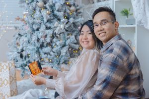 Pregnant couple sitting by Christmas tree holding ultrasound photo