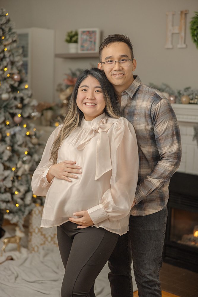 Pregnant couple standing together in front of Christmas tree home maternity session