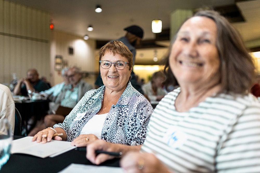 Two senior women smiling and socializing at business seminar table
