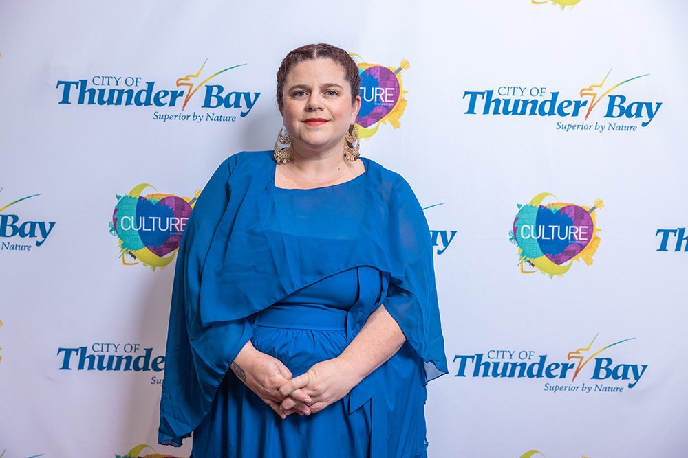 Woman in elegant blue formal dress with cape sleeves on red carpet step-and-repeat backdrop