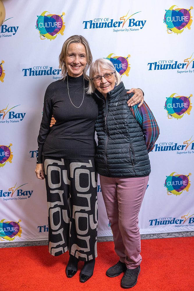 Two women embracing on red carpet, one in black patterned outfit, one in casual puffer vest